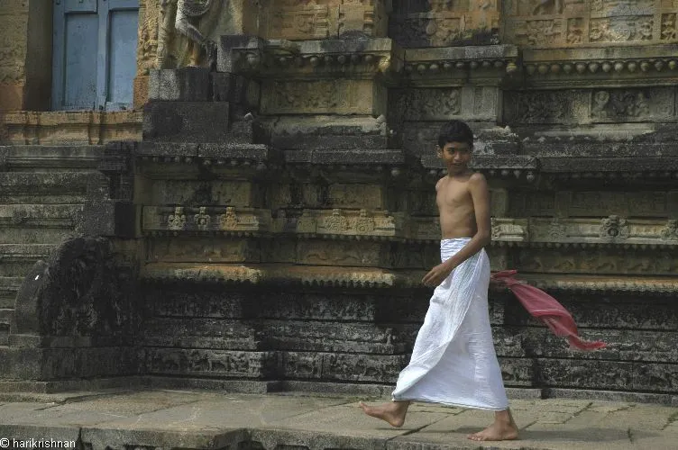 Boy in a white veshti, bare-chested, walking barefoot past a South Indian temple, the traditional temple dress for men