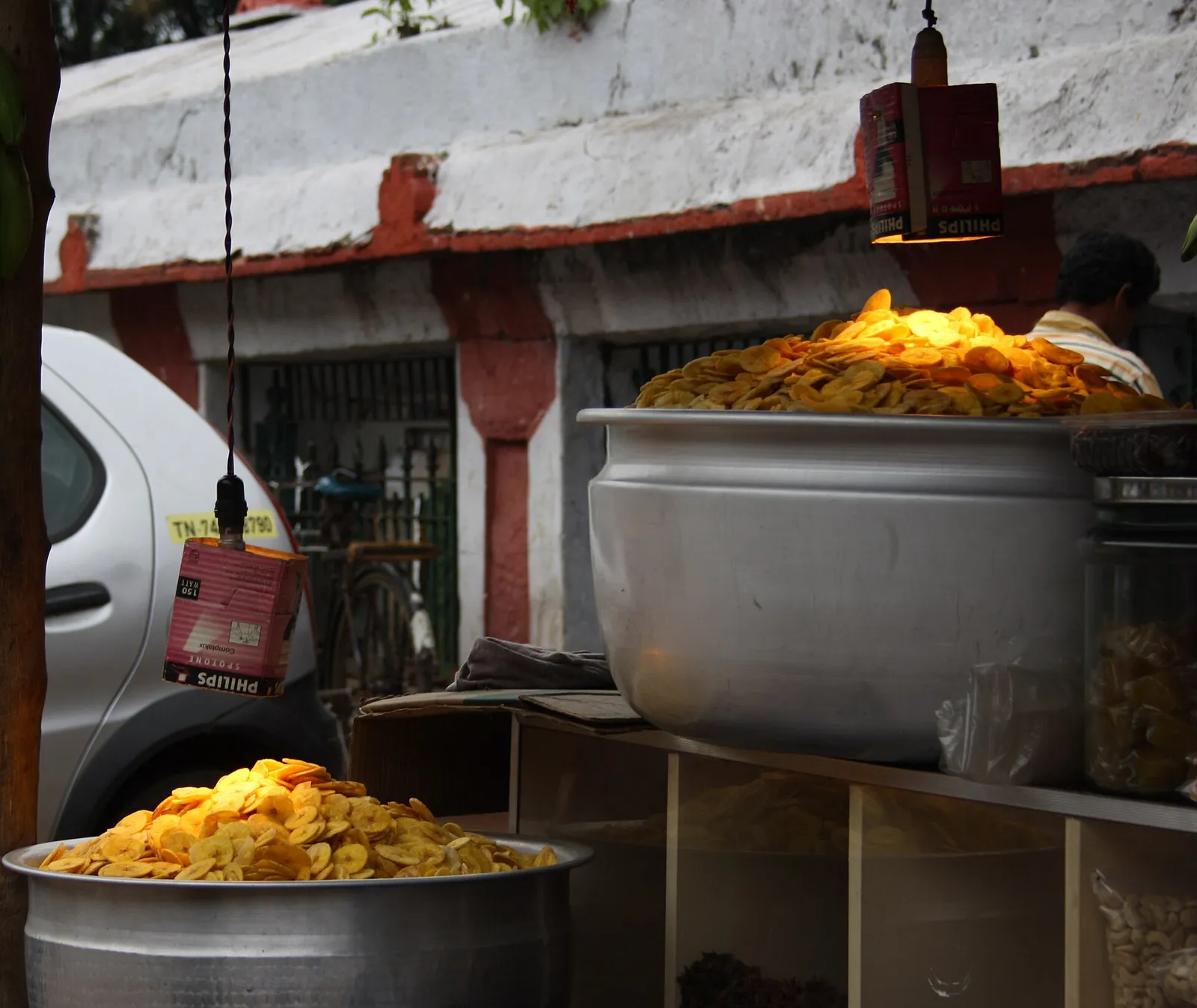 Freshly fried banana chips piled high in aluminium pots under hanging lamps at a roadside chips shop