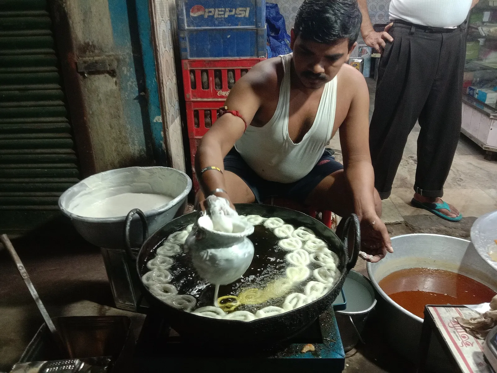 Jalebi being piped into hot oil at a local sweet shop, with a pot of sugar syrup alongside
